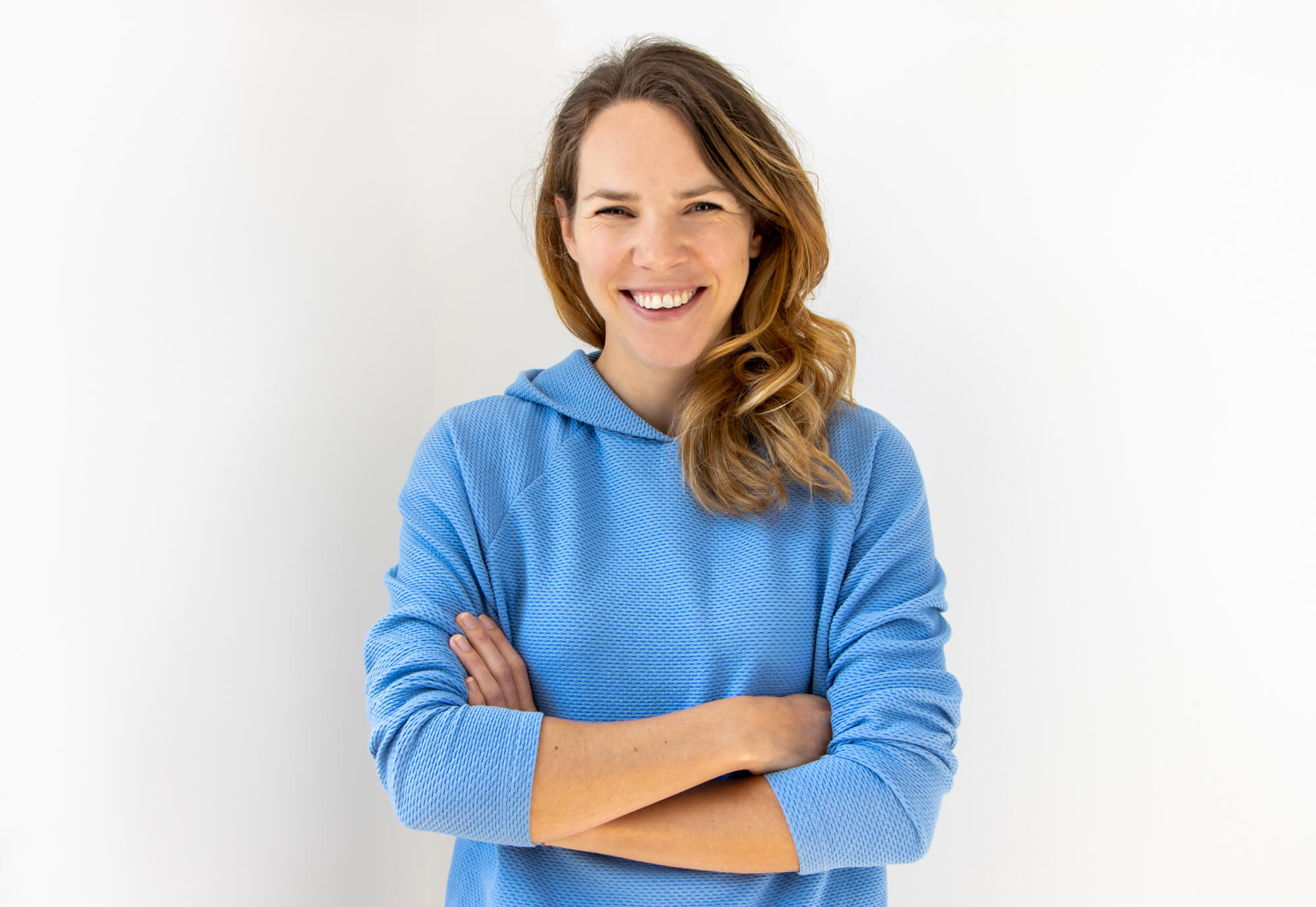 Eine lächelnde Person mit langen, welligen Haaren steht mit verschränkten Armen vor einem weißen Hintergrund.